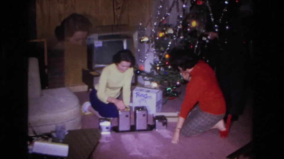 Two women examine Christmas gifts on the floor of a mid-1960s living room; a boxy period television, decorated tree with colored lights and tinsel, and branded cardboard boxes are visible.
