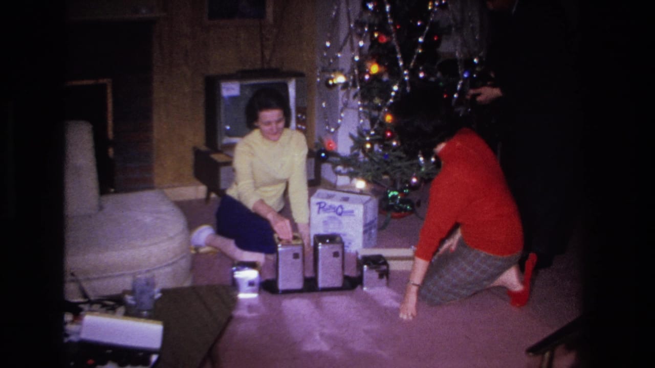 1966 8mm home movie: two women in yellow and red sweaters examine gifts beside a tinsel-lit Christmas tree in a wood-paneled living room in Hoboken, New Jersey.