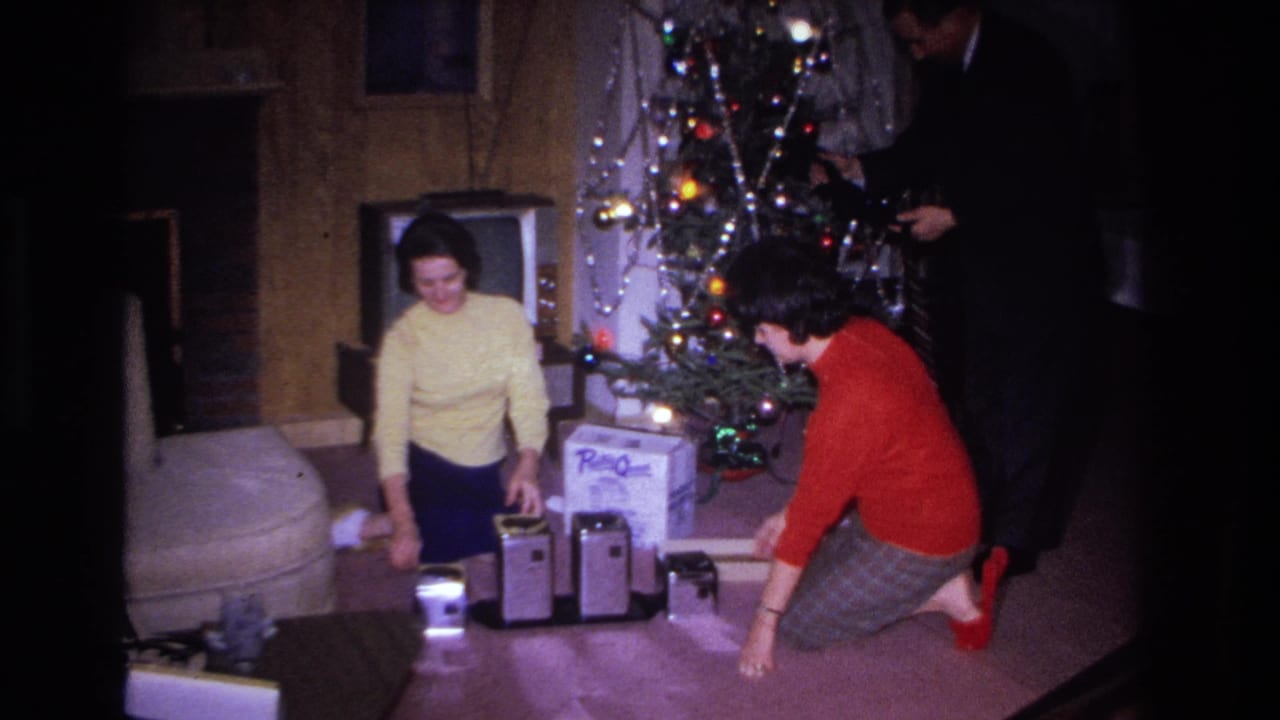 1966 8mm home movie: two women and a partially visible man in a dark jacket beside a Christmas tree with a RadioShack box and vintage electronics on the floor, Hoboken, NJ.