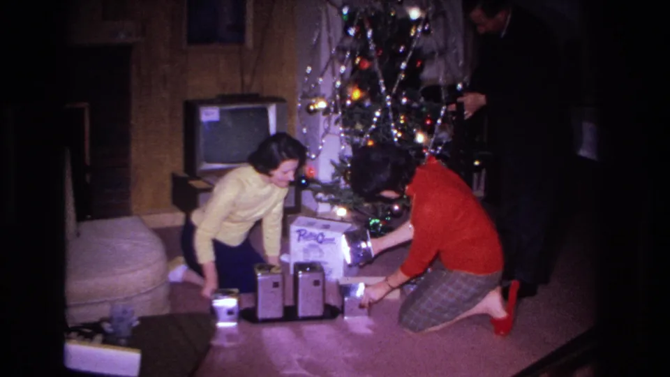 1966 8mm home movie: women in yellow and red sweaters crouch over floor gifts, with a CRT television and wood-paneled wall visible behind the decorated tree, Hoboken, NJ.