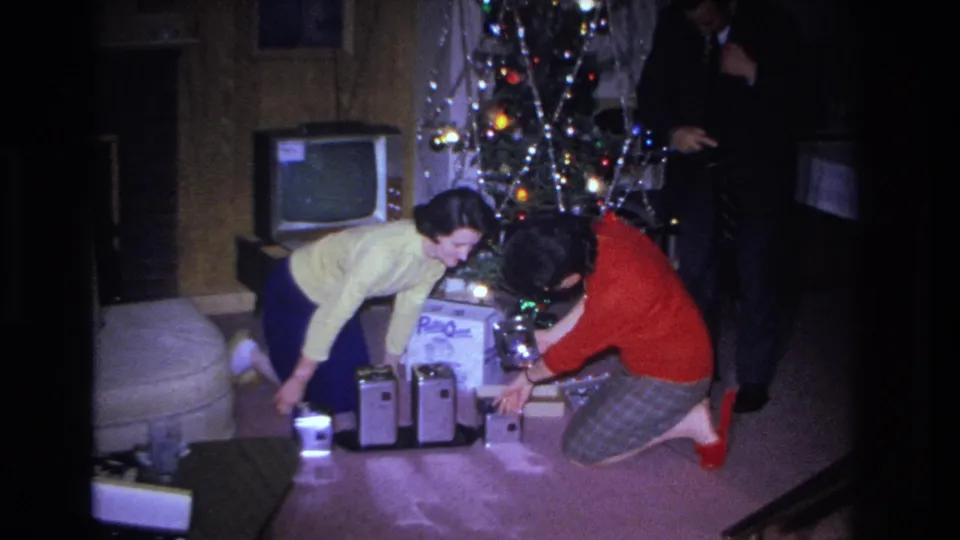 1966 8mm home movie: two women and a partially visible man gather around Christmas gifts in a wood-paneled living room with a CRT television visible in the background, Hoboken, NJ.