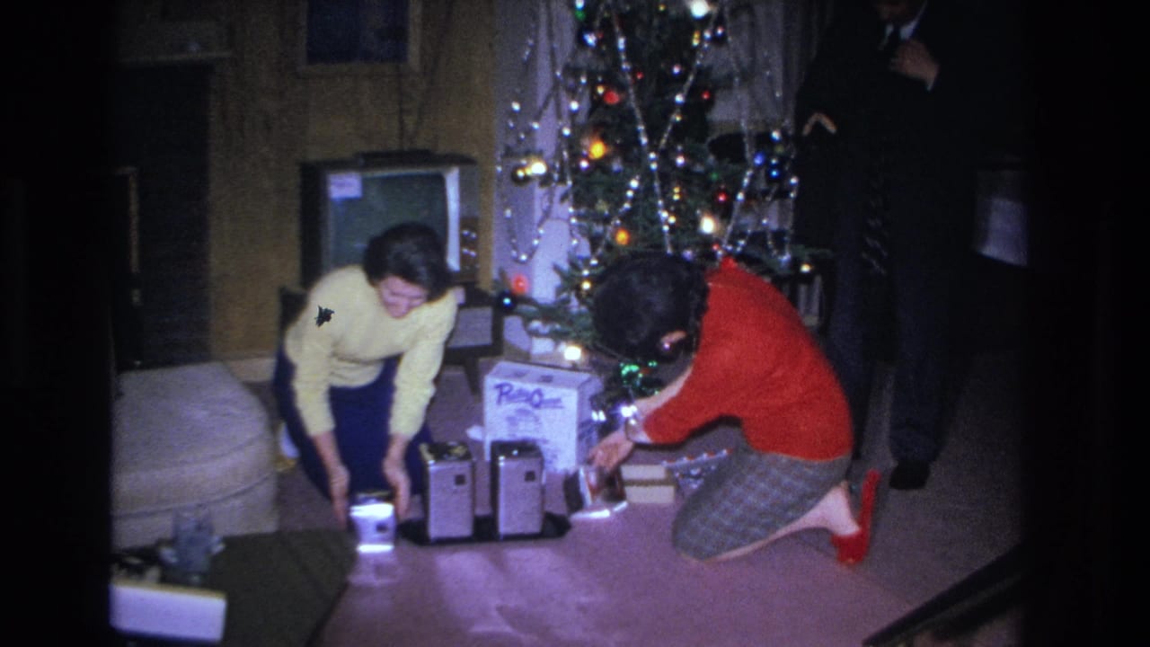 Two women in early-1960s clothing — yellow sweater and red top — opening gifts beside a tinsel-laden Christmas tree in a wood-paneled living room, circa 1966 Hoboken NJ.
