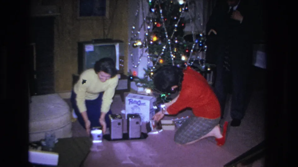 Two women in early-1960s clothing — yellow sweater and red top — opening gifts beside a tinsel-laden Christmas tree in a wood-paneled living room, circa 1966 Hoboken NJ.
