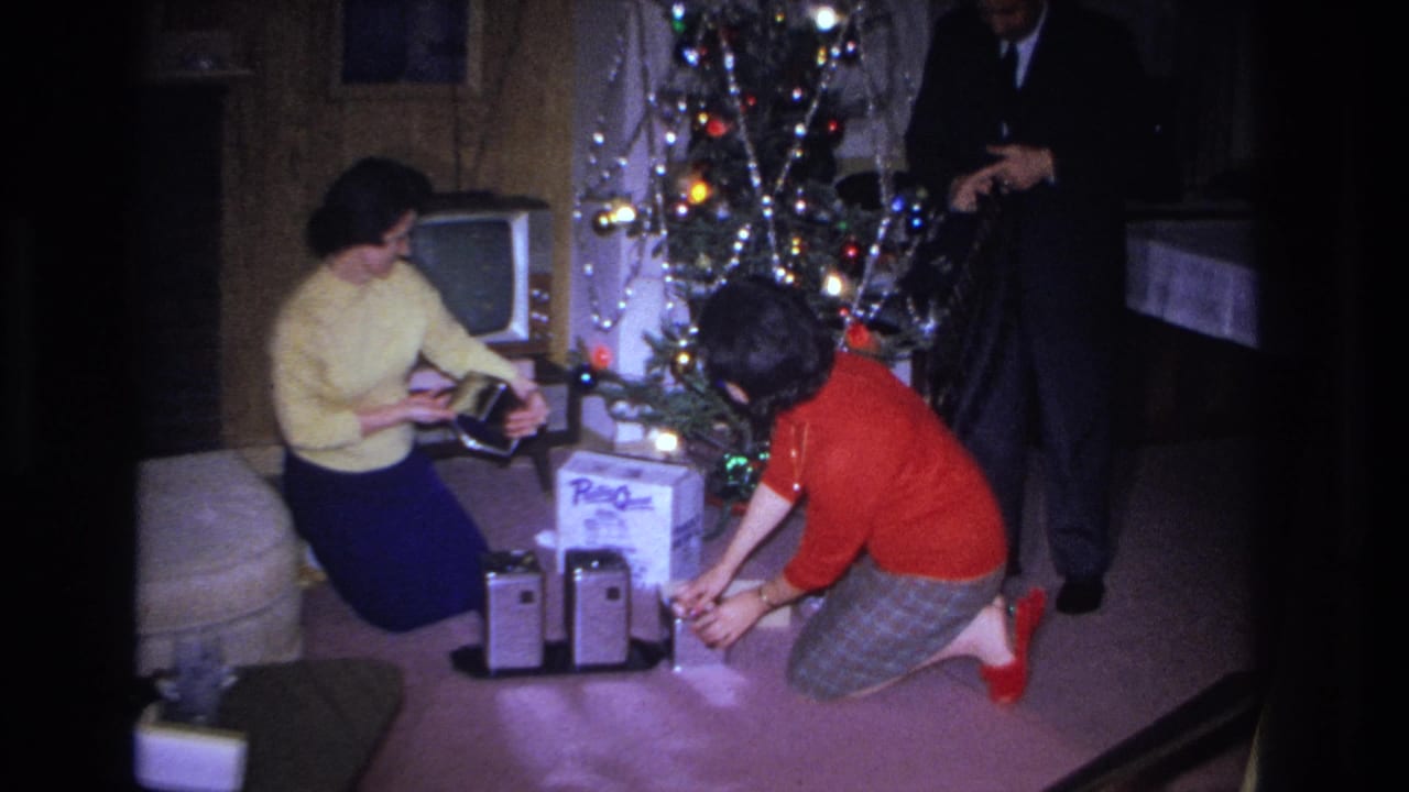 Two women examining a gift beside a lit Christmas tree; man in dark suit visible at right; television set in background.