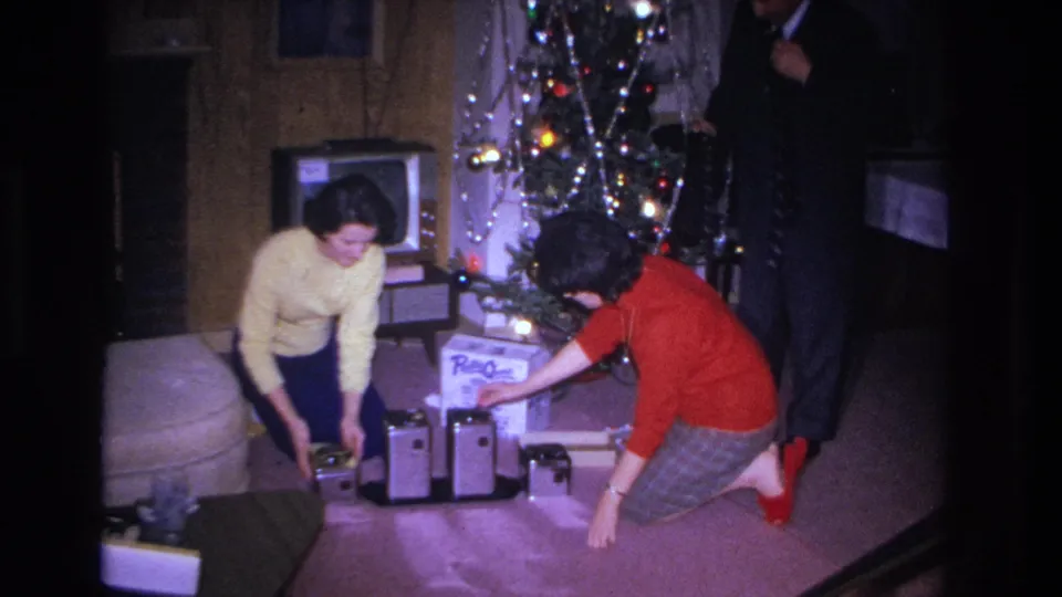 Wide view of two women in 1960s clothing reaching among gift boxes beneath a tinsel Christmas tree in a wood-paneled living room, circa 1966 Hoboken NJ.