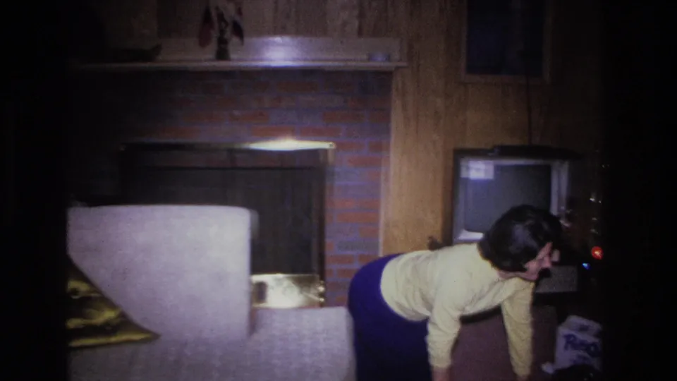 Woman with dark hair bending toward what appears to be an old television set in a 1960s wood-paneled living room; light upholstered sofa and brick fireplace visible in background.
