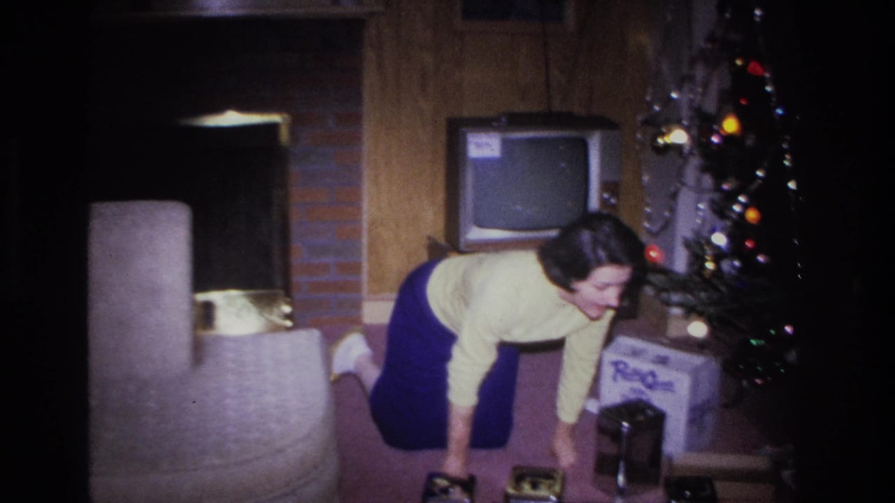 Woman in yellow sweater and blue skirt leans beside a lit Christmas tree in what appears to be a mid-1960s American living room, with a CRT television and wood-paneled wall visible.