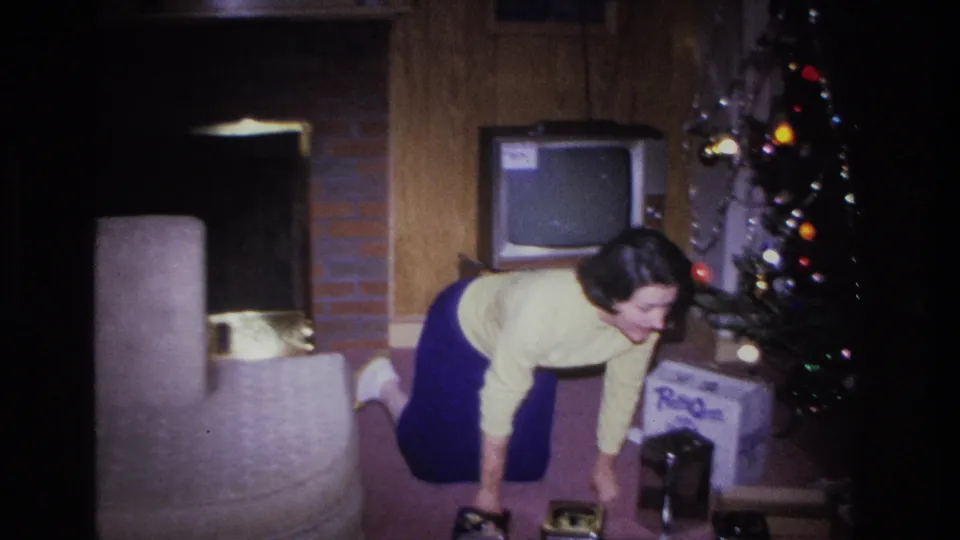 Woman in yellow sweater and blue skirt leans beside a lit Christmas tree in what appears to be a mid-1960s American living room, with a CRT television and wood-paneled wall visible.