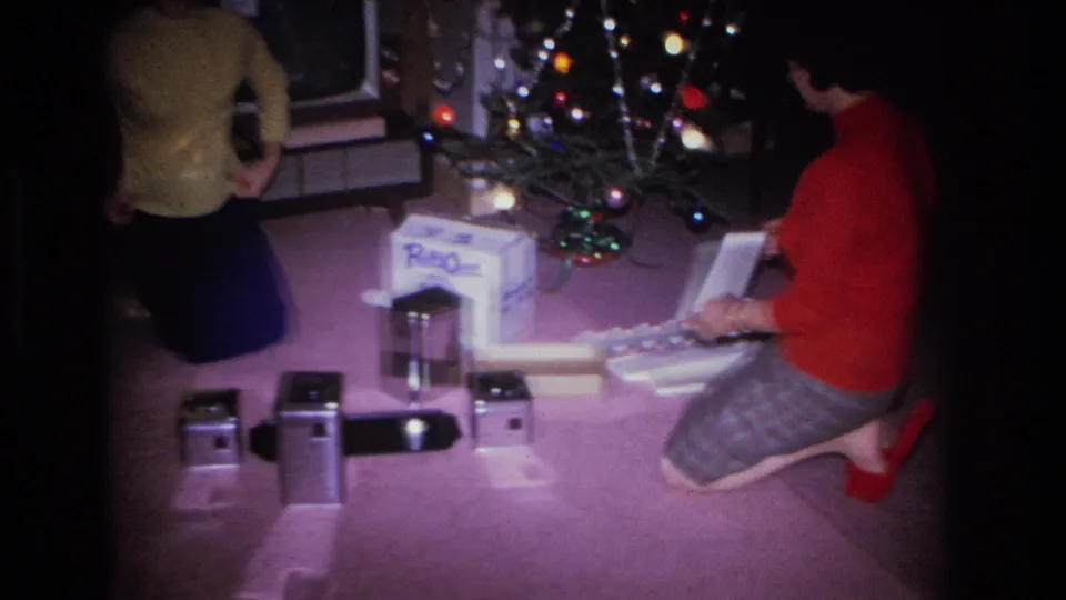 Two adults in a mid-1960s American living room: one in yellow sweater at frame left, one in red sweater kneeling among gifts beside a lit Christmas tree with colored bulbs.