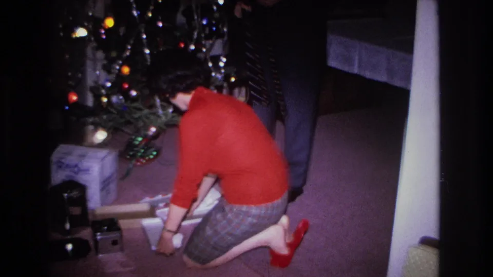 Person in red sweater kneels on a pink-carpeted floor handling a gift beside a decorated Christmas tree with colored bulbs, with small objects nearby and a second person partially visible at right.