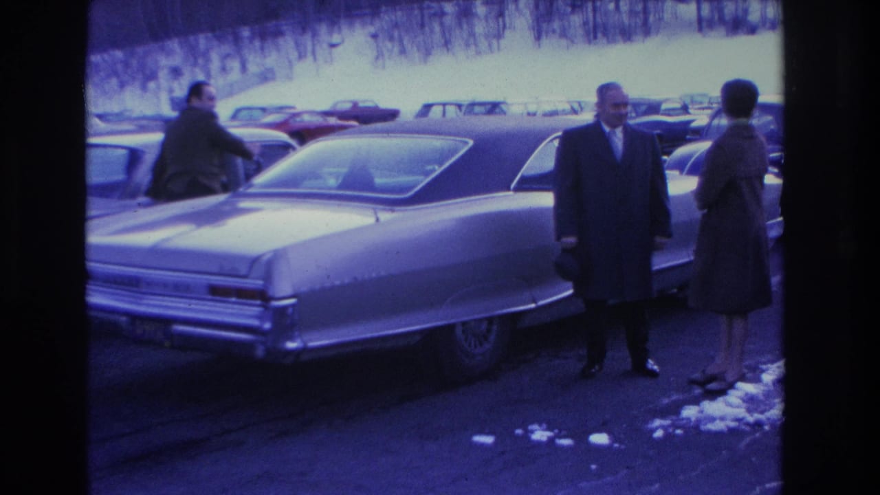 Man in dark overcoat and woman in dark coat stand beside a large American sedan in a snowy parking lot; Hoboken, New Jersey, 1966.