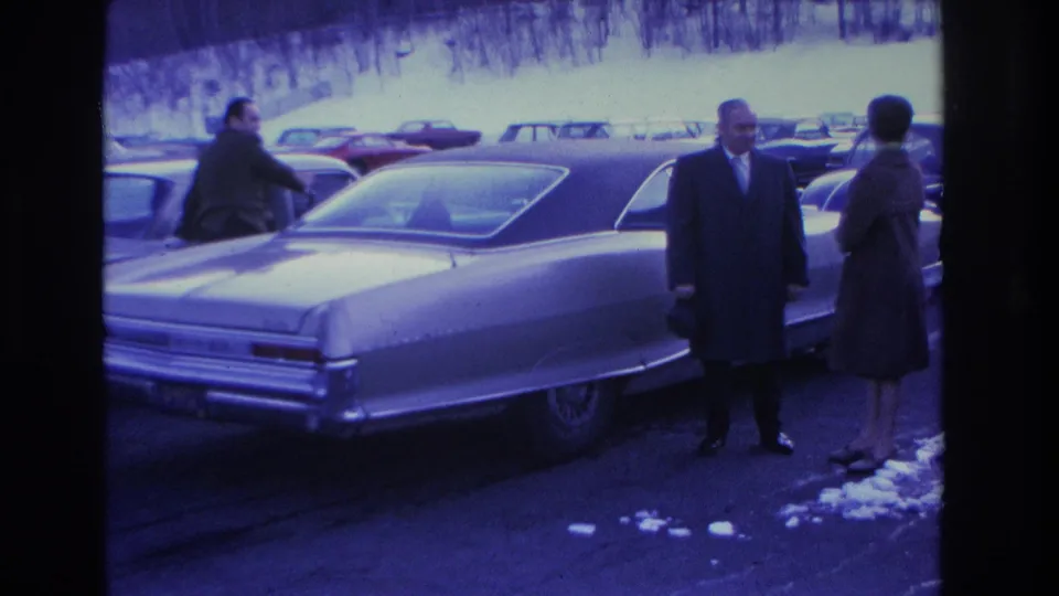Man in dark overcoat and woman in dark coat stand beside a large American sedan in a snowy parking lot; Hoboken, New Jersey, 1966.
