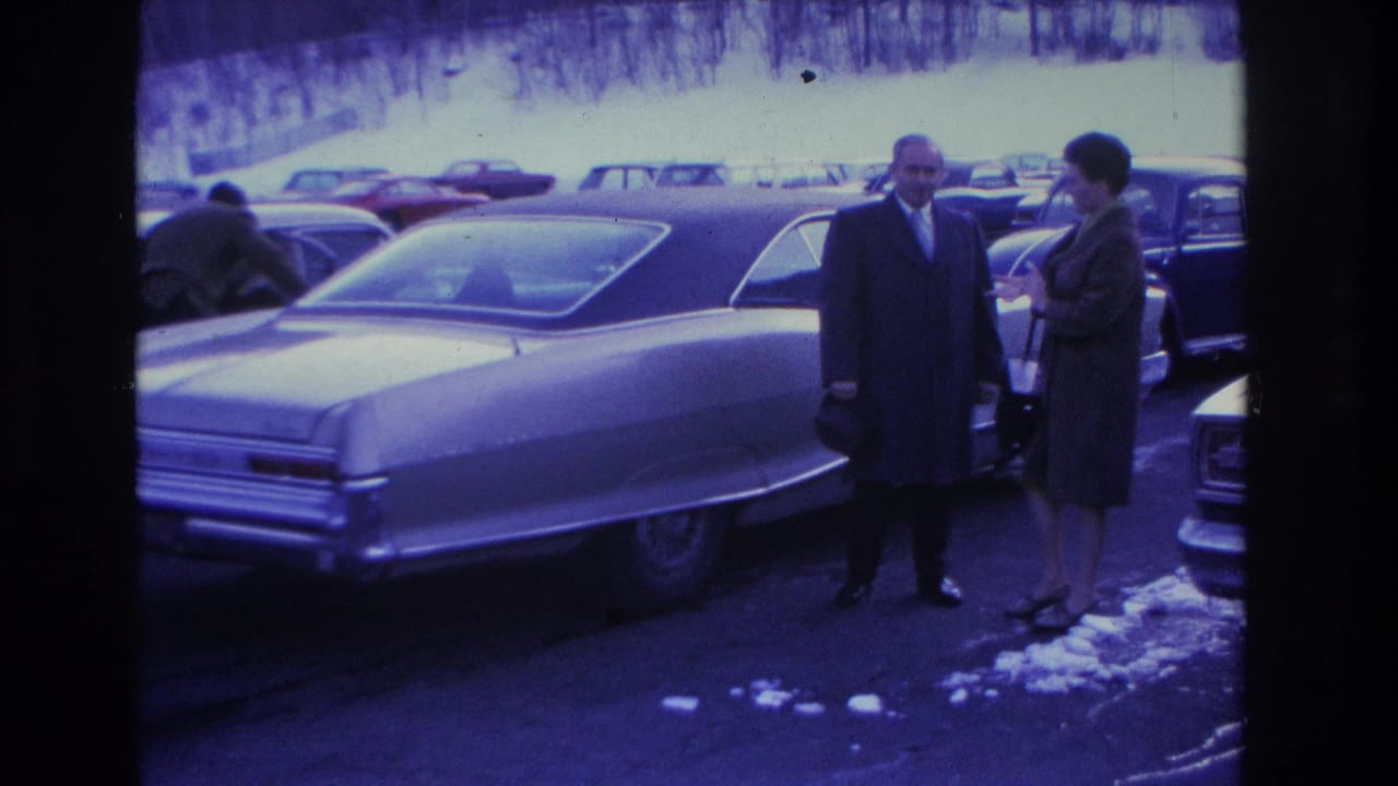Man in overcoat and woman in dark coat beside a large sedan; background figure and row of 1960s cars in snowy parking lot.