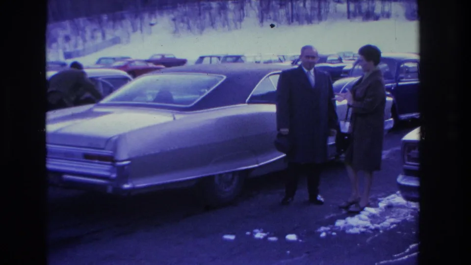 Man in dark overcoat and woman in dark coat beside a large 1960s American sedan; a third person visible in background; Hoboken, New Jersey, 1966.