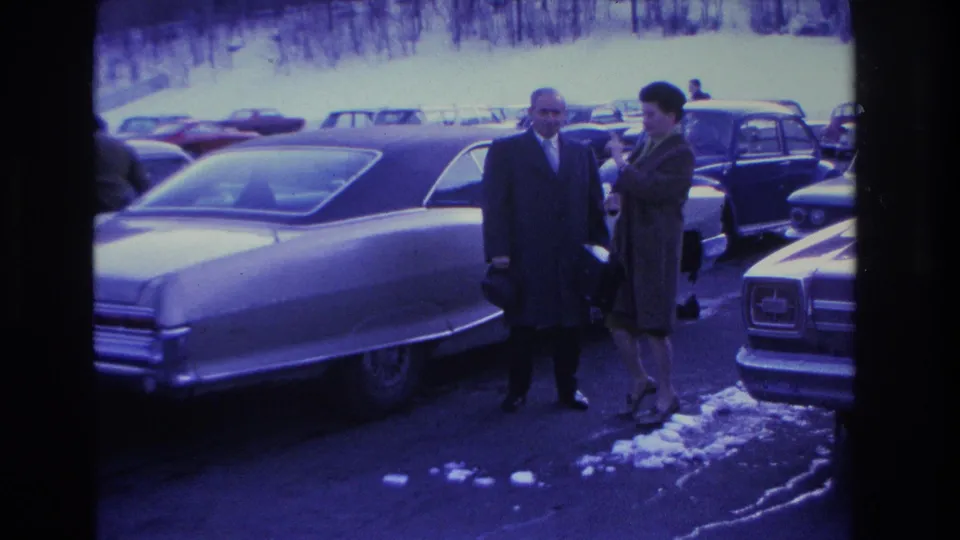 Man in dark overcoat and woman in dark coat begin posing for the camera in a snowy parking lot full of 1960s American automobiles; Hoboken, New Jersey, 1966.