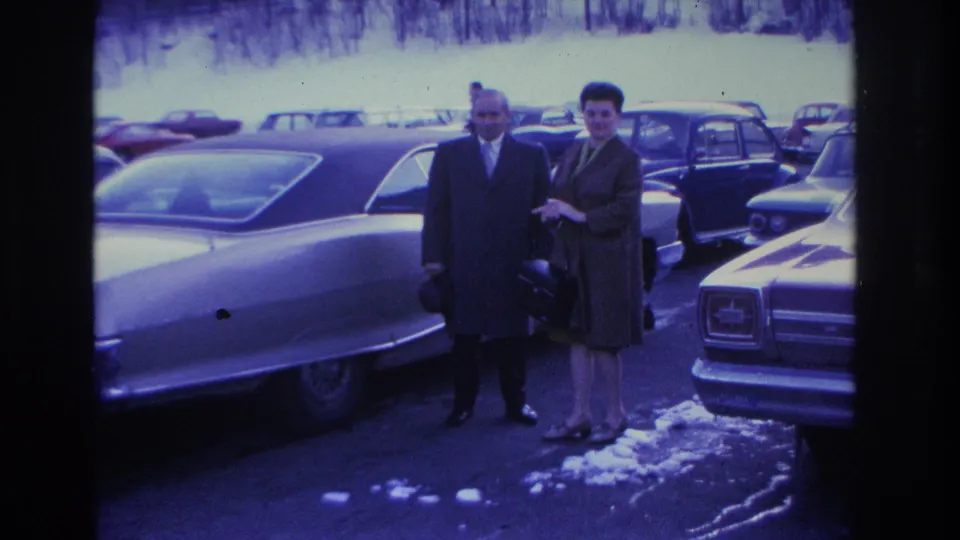 Man and woman in formal winter dress face the camera in a snowy parking lot beside a large American sedan; the woman appears to smile; Hoboken, New Jersey, 1966.