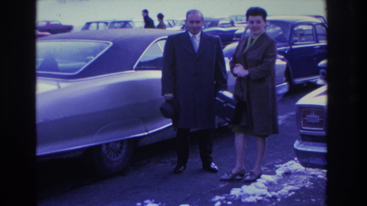 Couple in formal winter coats pose in a snowy parking lot amid 1960s American sedans, Hoboken, New Jersey, circa 1966; blue color cast from aged 8mm film.
