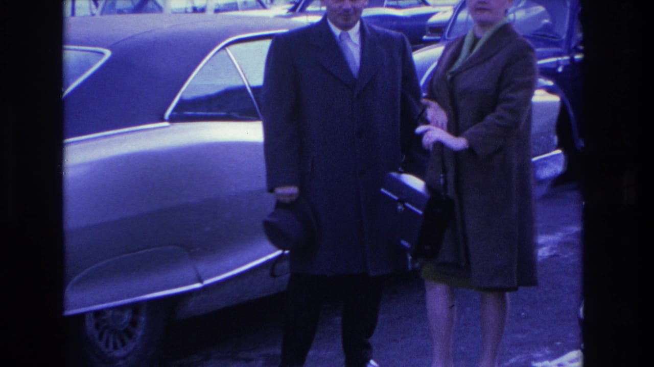 Mid-shot of a couple in formal winter coats beside a 1960s American sedan; strong blue-violet color cast from aged 8mm film, circa 1966 Hoboken, New Jersey.