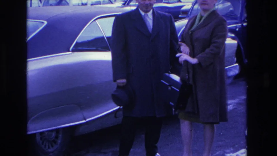 Mid-shot of a couple in formal winter coats beside a 1960s American sedan; strong blue-violet color cast from aged 8mm film, circa 1966 Hoboken, New Jersey.