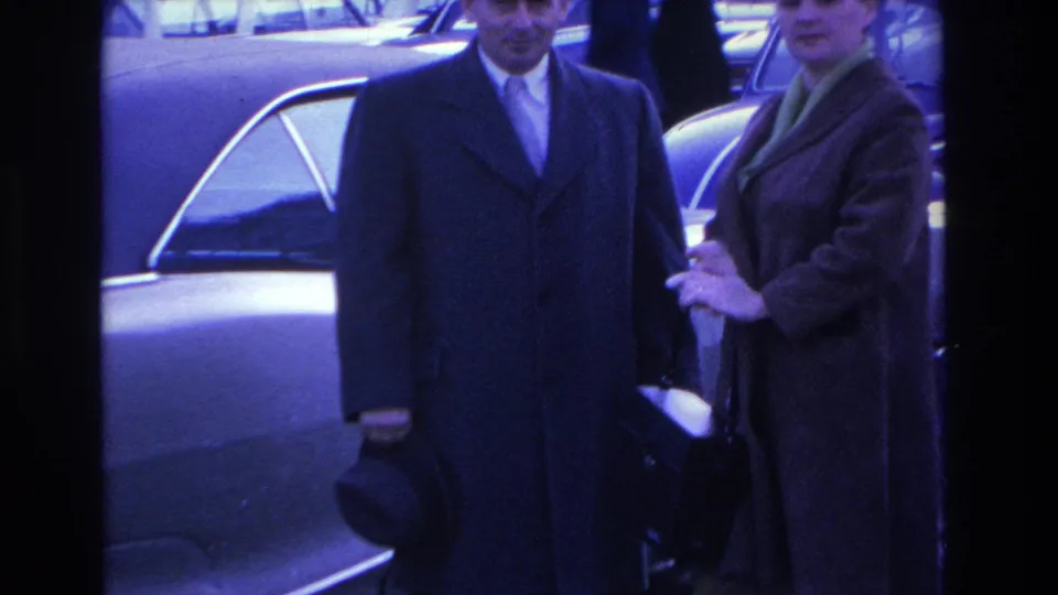 Man in dark overcoat holding a fedora and woman in tweed coat pause in a parking lot; candid posing moment on 8mm, circa 1966 Hoboken, New Jersey.