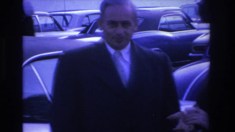 Close-up of a man in dark overcoat and tie turning smiling toward the camera; motion blur from handheld 8mm, circa 1966 Hoboken, New Jersey.