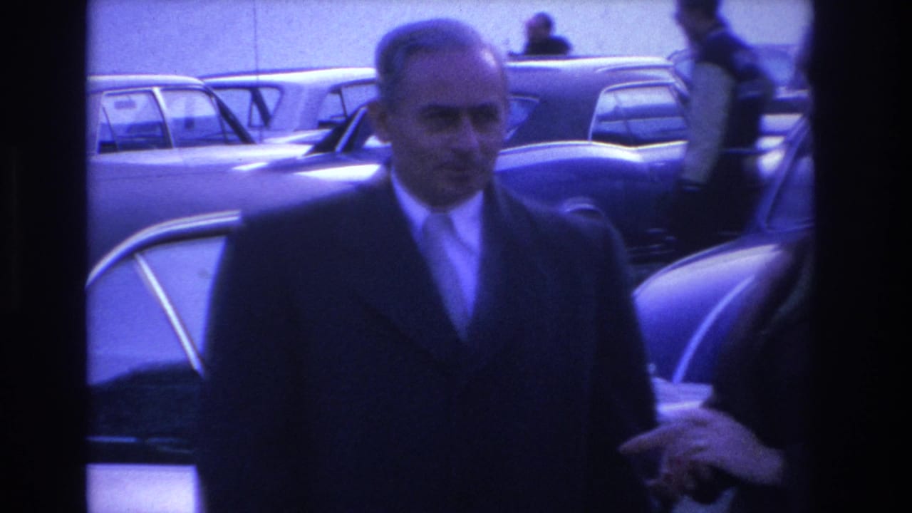 Close-up of a man in a dark overcoat and tie among mid-1960s American sedans in a parking lot, from an 8mm home movie circa 1966.