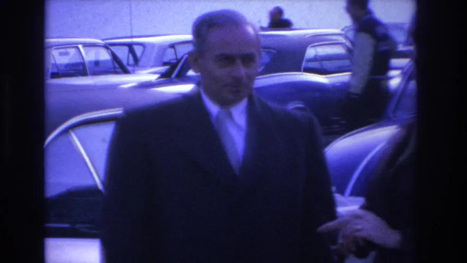 Close-up of a man in a dark overcoat and tie among mid-1960s American sedans in a parking lot, from an 8mm home movie circa 1966.