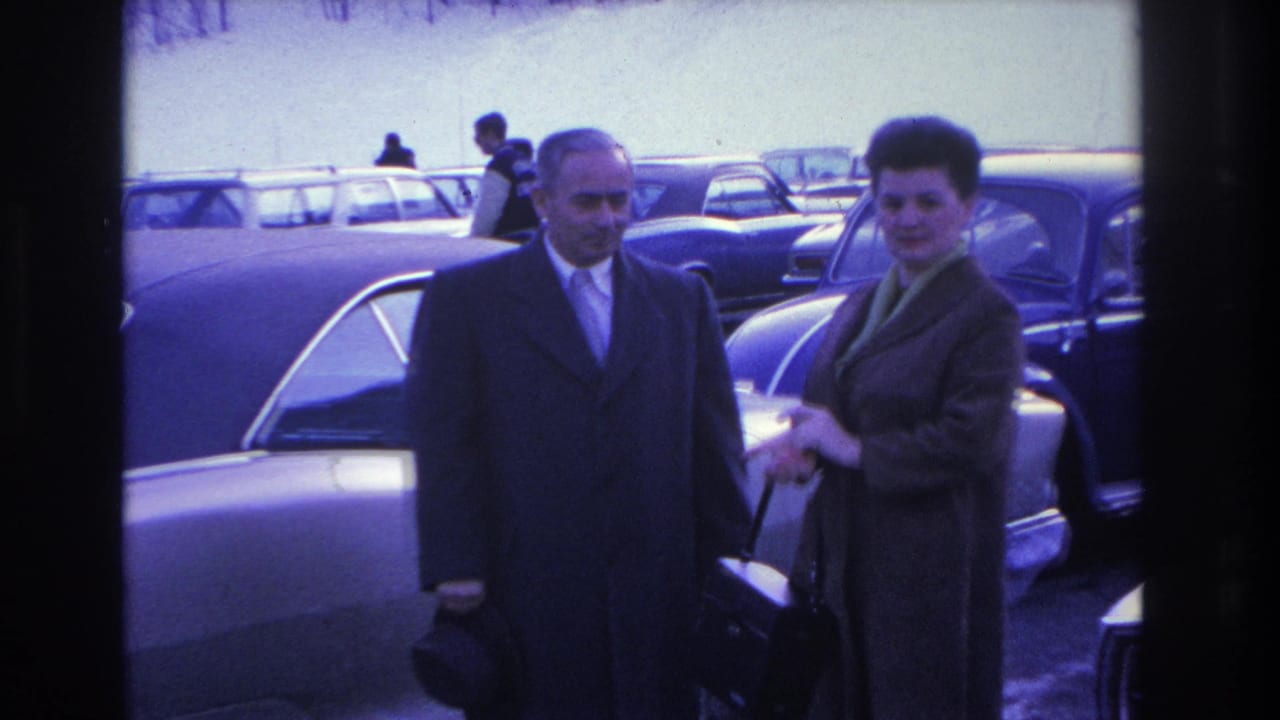 Man in dark overcoat and woman in long coat stand together in a parking lot among classic American cars.