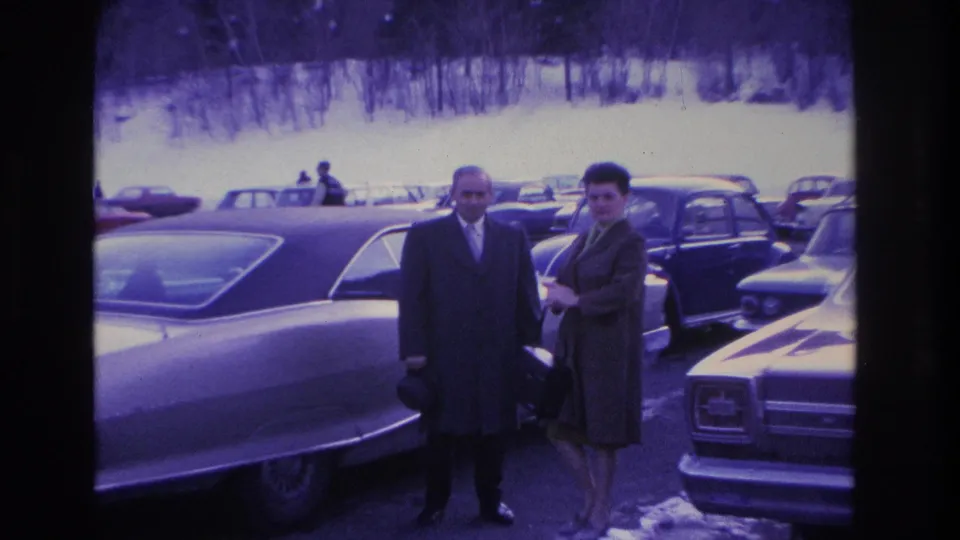 Wider view of a couple in winter coats in a packed mid-1960s parking lot with a snow-covered hillside and bare deciduous trees behind, from an 8mm home movie circa 1966.
