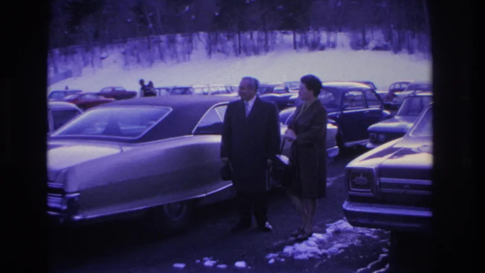 Couple in winter coats beside a large American sedan with chrome trim in a snow-dusted parking lot, from an 8mm home movie attributed to Hoboken, New Jersey, circa 1966.