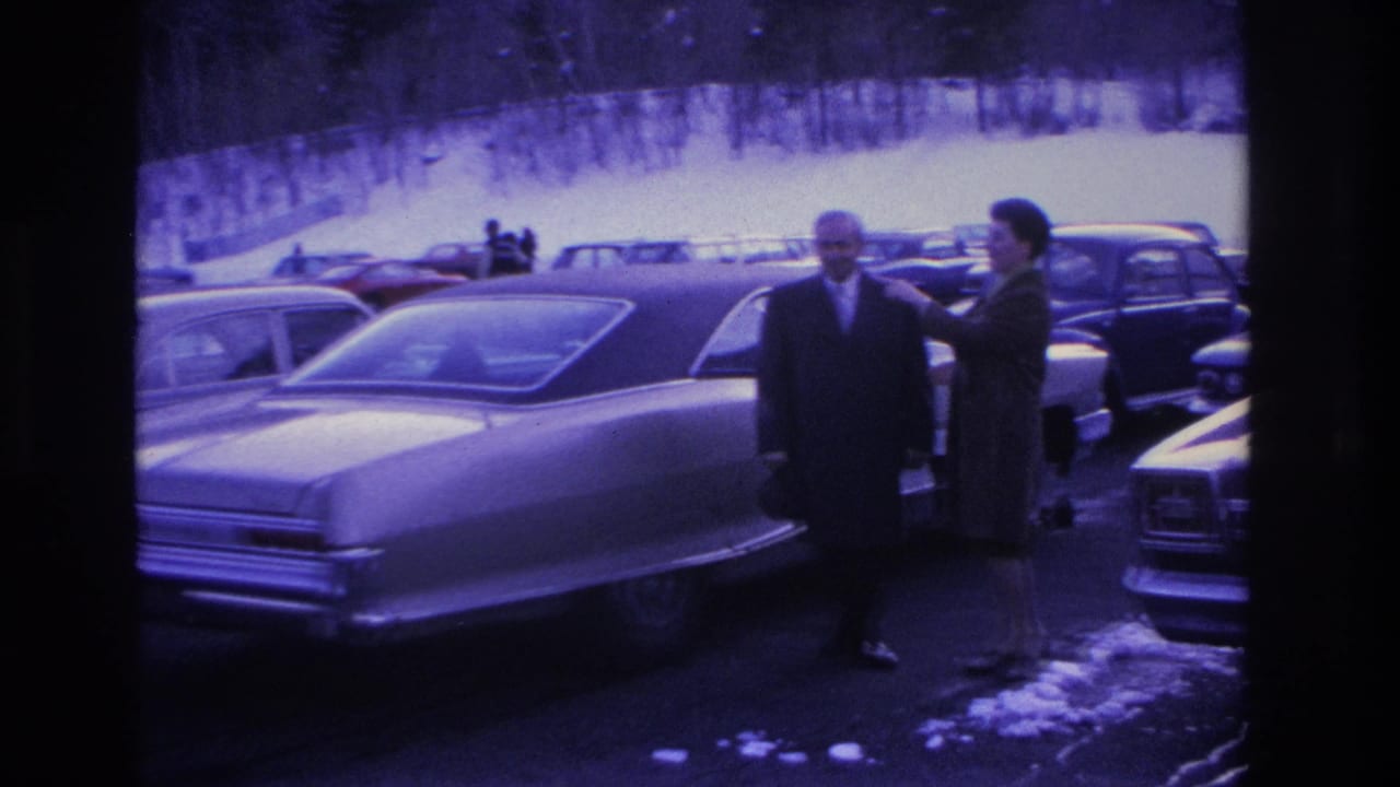 Man in dark suit and woman in dark coat beside a mid-1960s full-size American sedan in a snowy parking lot; Hoboken, NJ, 1966; pronounced purple-blue 8mm film shift.