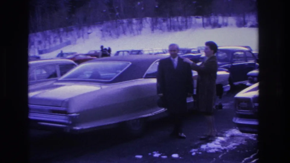 Man in dark suit and woman in dark coat beside a mid-1960s full-size American sedan in a snowy parking lot; Hoboken, NJ, 1966; pronounced purple-blue 8mm film shift.