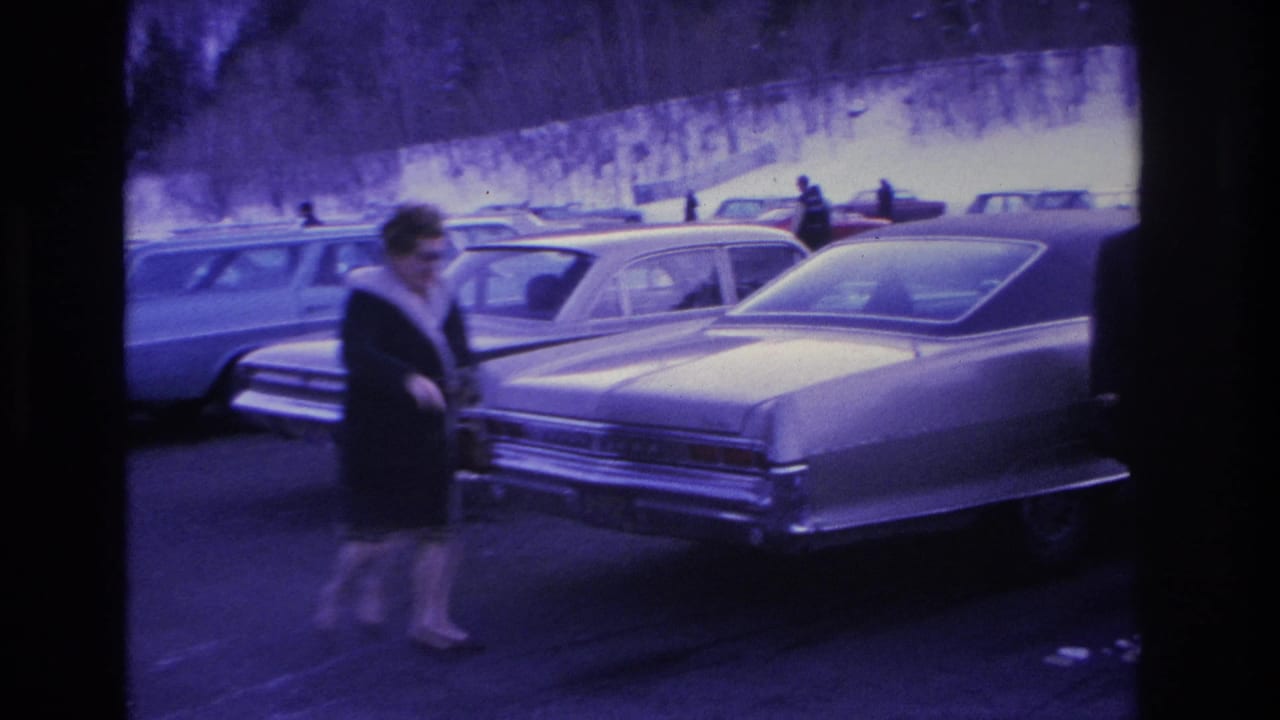 Woman in dark coat walking through a snowy parking lot past mid-1960s American sedans; 8mm footage with heavy purple-blue film shift, Hoboken, NJ, circa 1966.