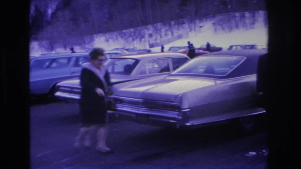 Woman in dark coat walking through a snowy parking lot past mid-1960s American sedans; 8mm footage with heavy purple-blue film shift, Hoboken, NJ, circa 1966.