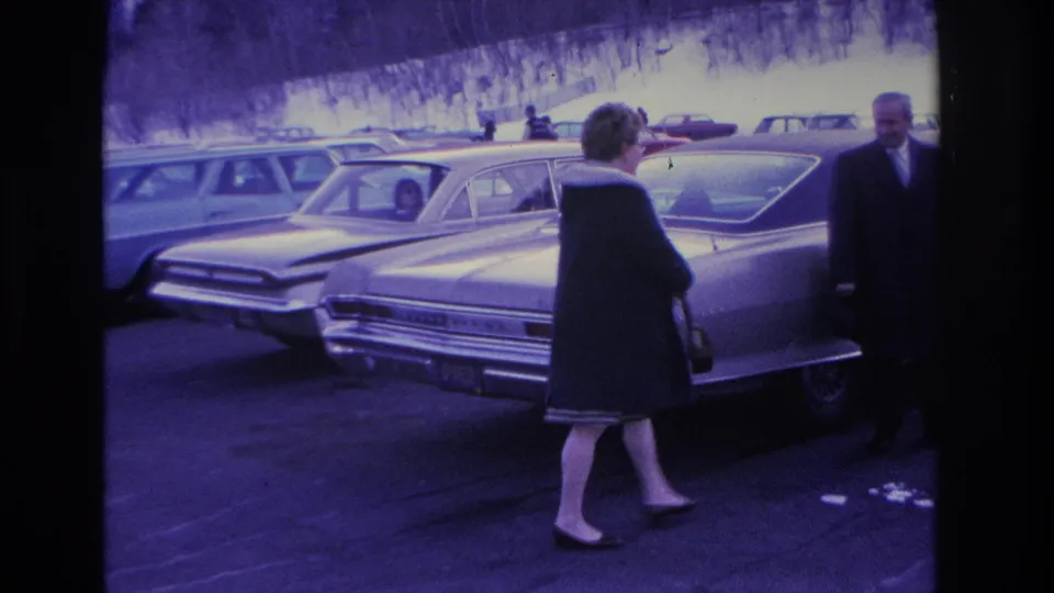 Woman in dark coat walking toward camera while a suited man stands in background among 1960s American sedans in a snowy lot; 8mm footage with purple-blue film tint, circa 1966.