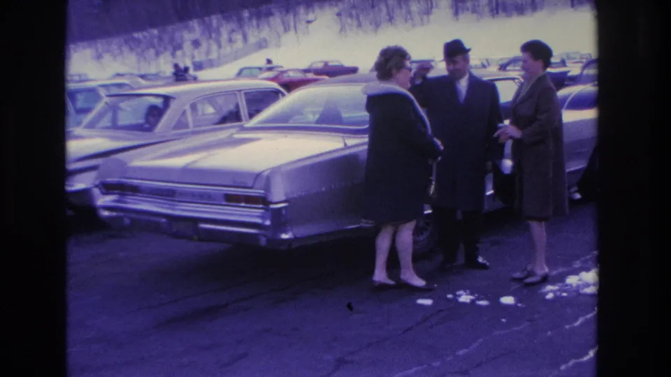 Two women and a hatted, suited man in conversation beside a mid-1960s American sedan in a snowy parking lot; 8mm home movie, Hoboken, NJ, circa 1966.