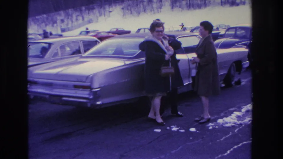Two women in 1960s winter coats and handbags beside a large mid-decade American sedan in a winter parking lot, Hoboken, New Jersey area, circa 1966.