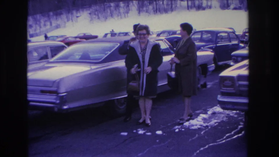 Two women in mid-1960s winter coats — one wearing glasses — near the rear of a large American sedan in a snow-patched parking lot, Hoboken, New Jersey area, circa 1966.