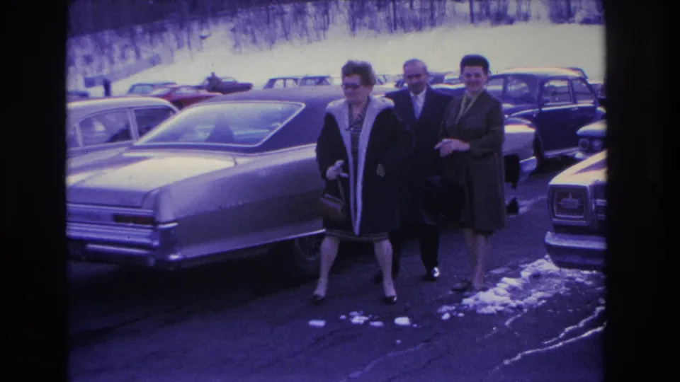 A man and two women in mid-1960s winter dress walking beside a large American sedan in a snow-patched parking lot, Hoboken, New Jersey area, circa 1966.