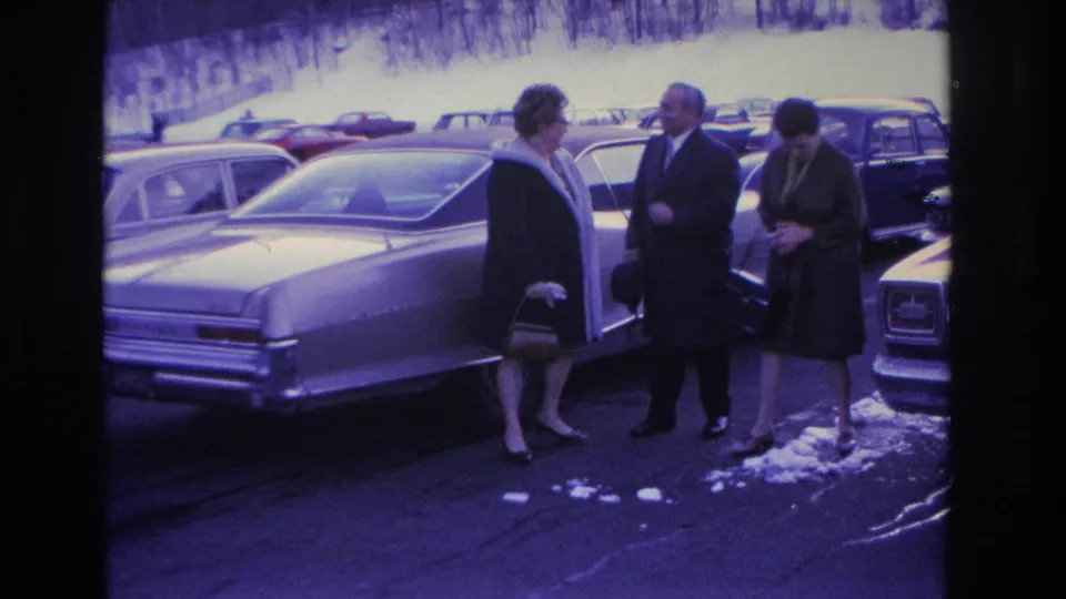 A man and two women in 1960s winter dress conversing beside the rear of a large American sedan in a snow-patched parking lot, Hoboken, New Jersey area, circa 1966.