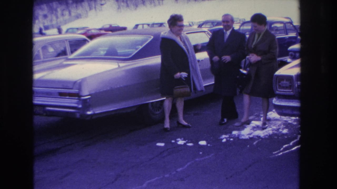 Three adults in winter coats converse beside a large American sedan in a snowy parking lot, likely Hoboken, New Jersey, circa 1966; purple color cast from aged 8mm film.
