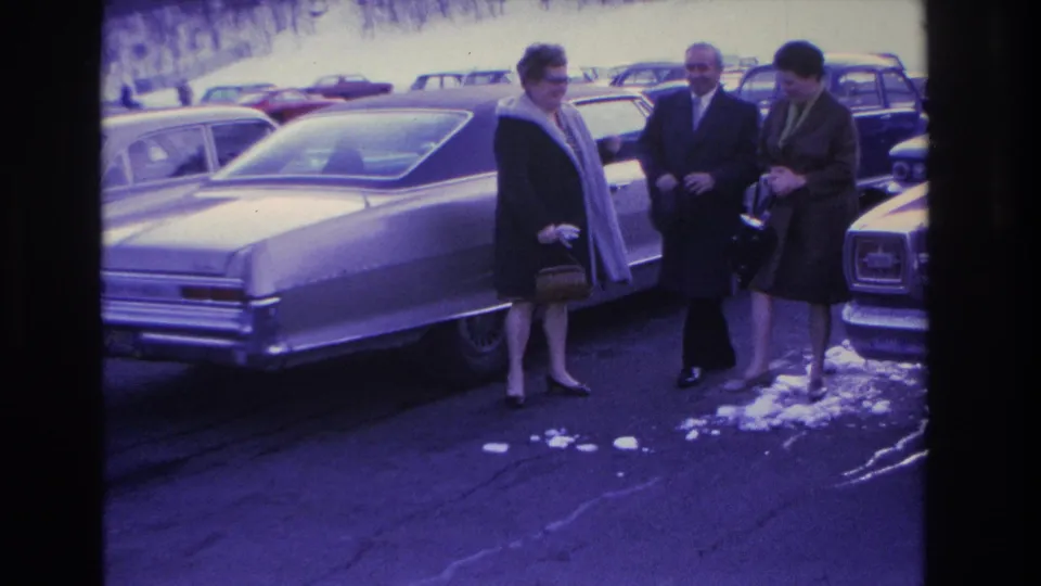 Three adults in winter coats converse beside a large American sedan in a snowy parking lot, likely Hoboken, New Jersey, circa 1966; purple color cast from aged 8mm film.