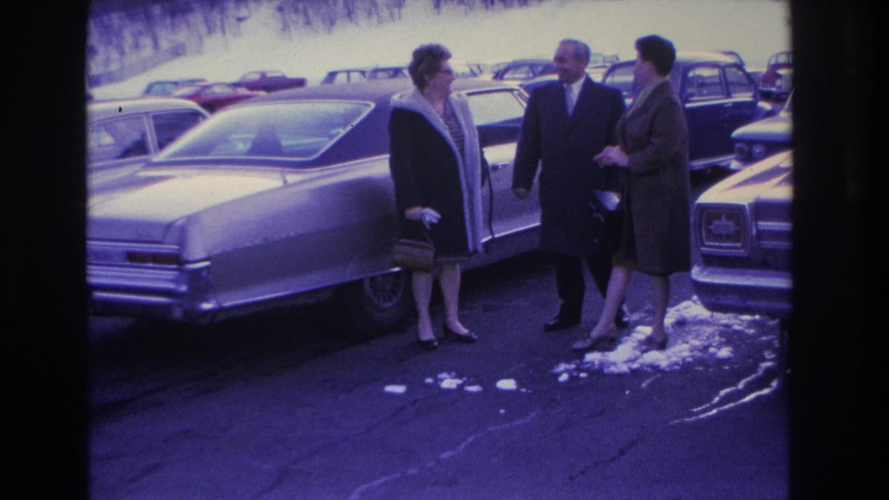 Three adults conversing in a snowy parking lot, large American sedan prominent in foreground, red car visible in background.
