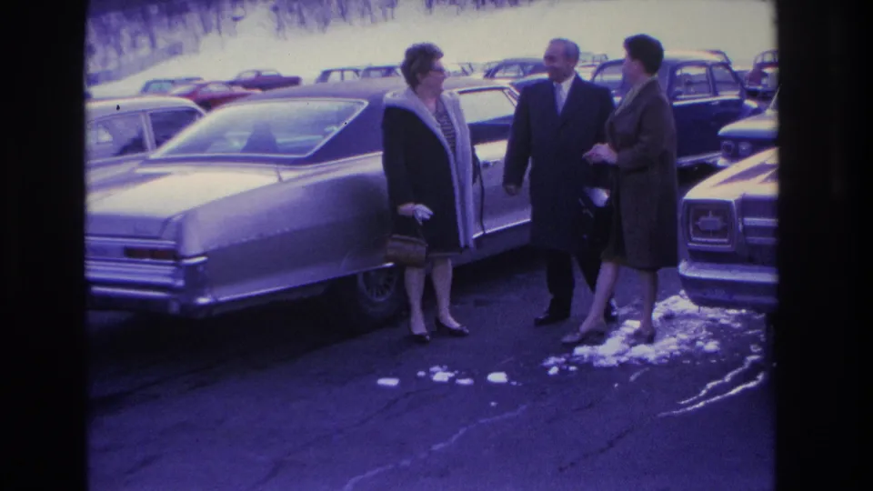 Three adults mid-conversation beside a large 1960s American sedan in a snowy parking lot, Hoboken, New Jersey area, circa 1966; purple-shifted aged 8mm film with visible grain.
