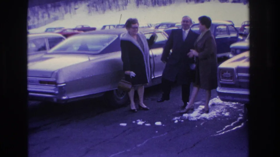 Three adults repositioning in a snowy parking lot beside a large 1960s sedan, circa 1966 Hoboken, New Jersey; purple cast and grain from aged 8mm reversal stock.