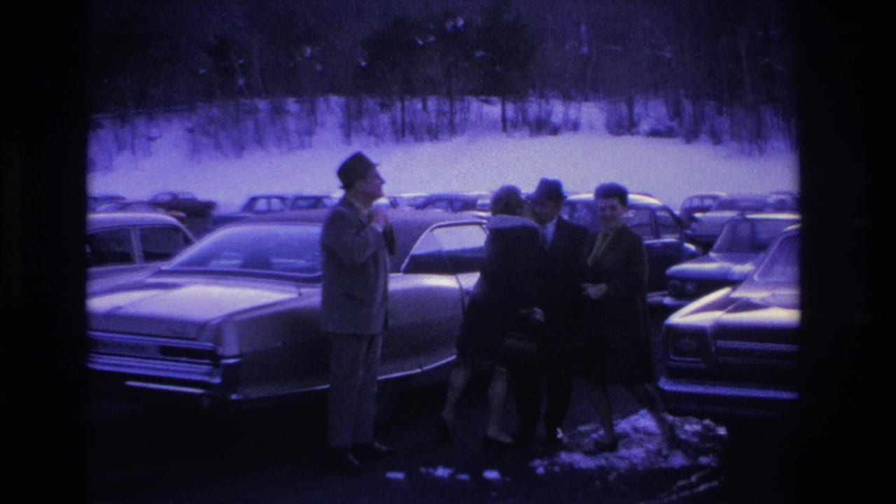 Four adults in winter overcoats and hats gathered beside a classic mid-1960s sedan in a snow-covered parking lot, Hoboken, New Jersey area, circa 1966; purple-blue film tint.