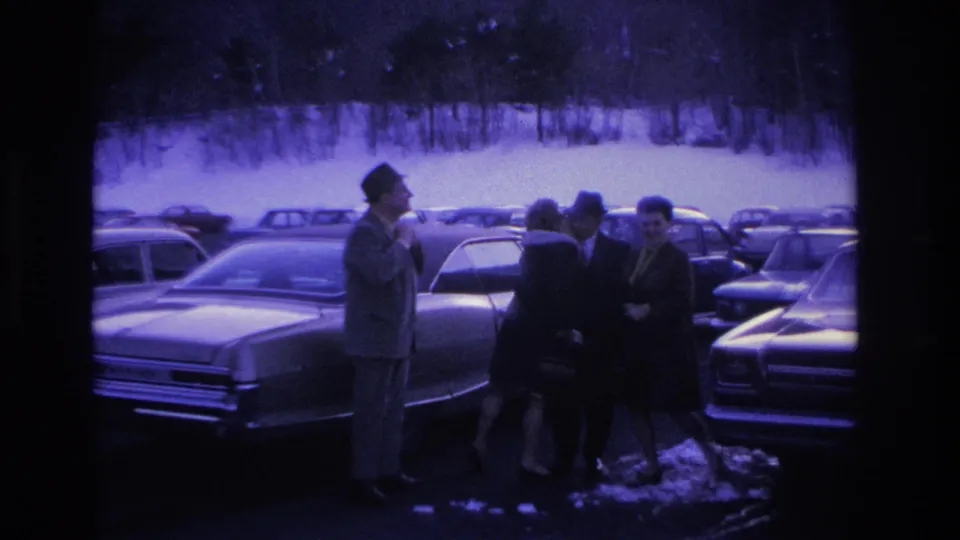 Four adults in winter overcoats and hats gathered beside a classic mid-1960s sedan in a snow-covered parking lot, Hoboken, New Jersey area, circa 1966; purple-blue film tint.