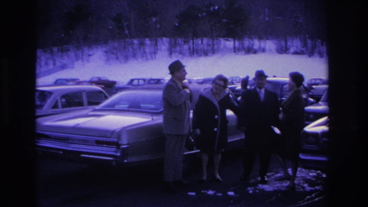 Group of four adults in winter dress conversing beside a large American sedan in a snow-covered parking lot.