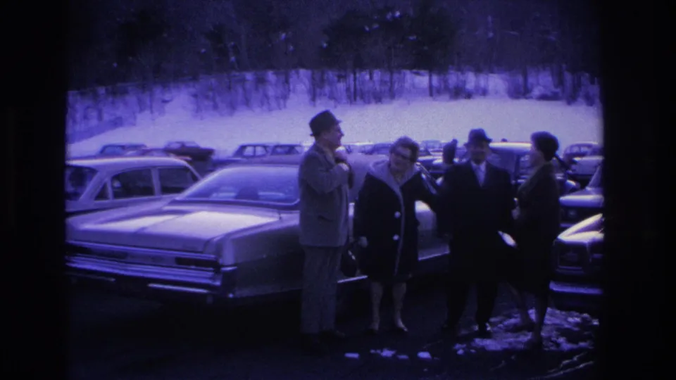 Four adults—two men in fedoras and overcoats, two women—conversing next to a mid-1960s sedan in a snowy parking lot, with rows of period vehicles visible behind them.
