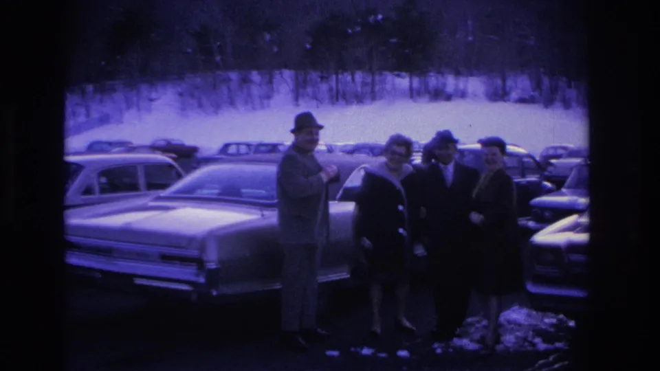 Four adults gathered near a classic American sedan in a snowy parking lot, circa 1966, with additional period vehicles and bare winter trees filling the background.